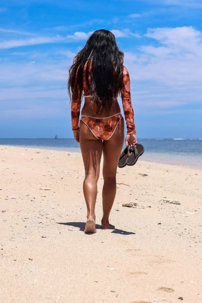 Woman in a red bikini and patterned shirt walking on a beach with a blue sky holding Olukai sandals